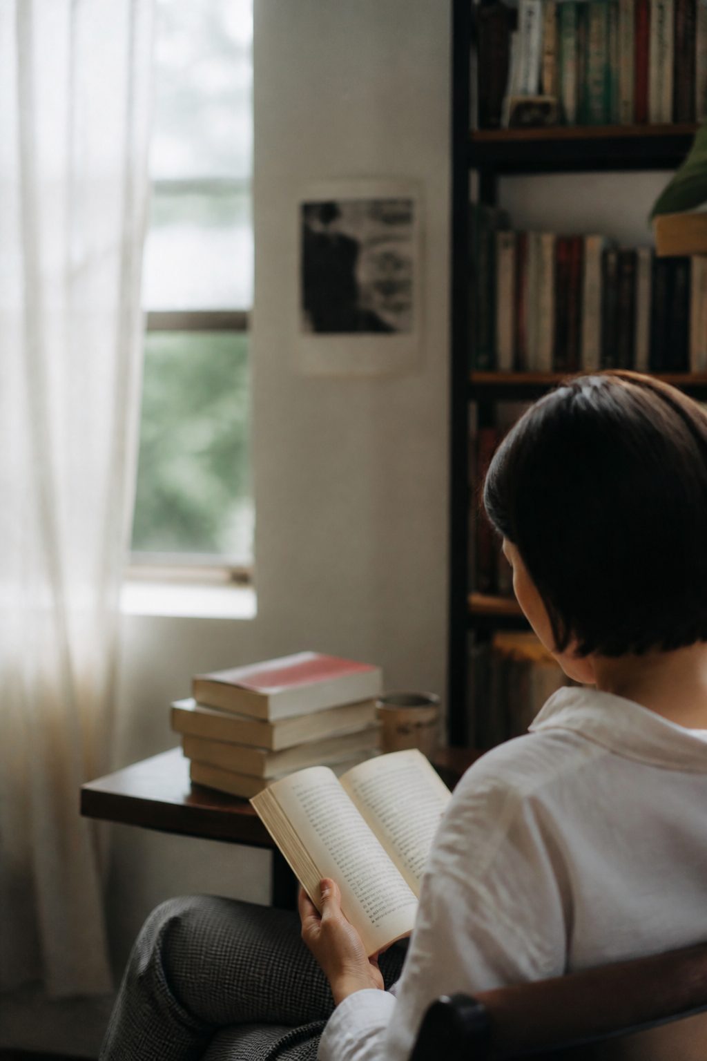 Una mesa de madera junto a una ventana con luz de tarde, sobre ella un libro abierto boca abajo, un cuaderno con notas fechadas y un lápiz; al fondo, una estantería con libros de distintos tamaños.